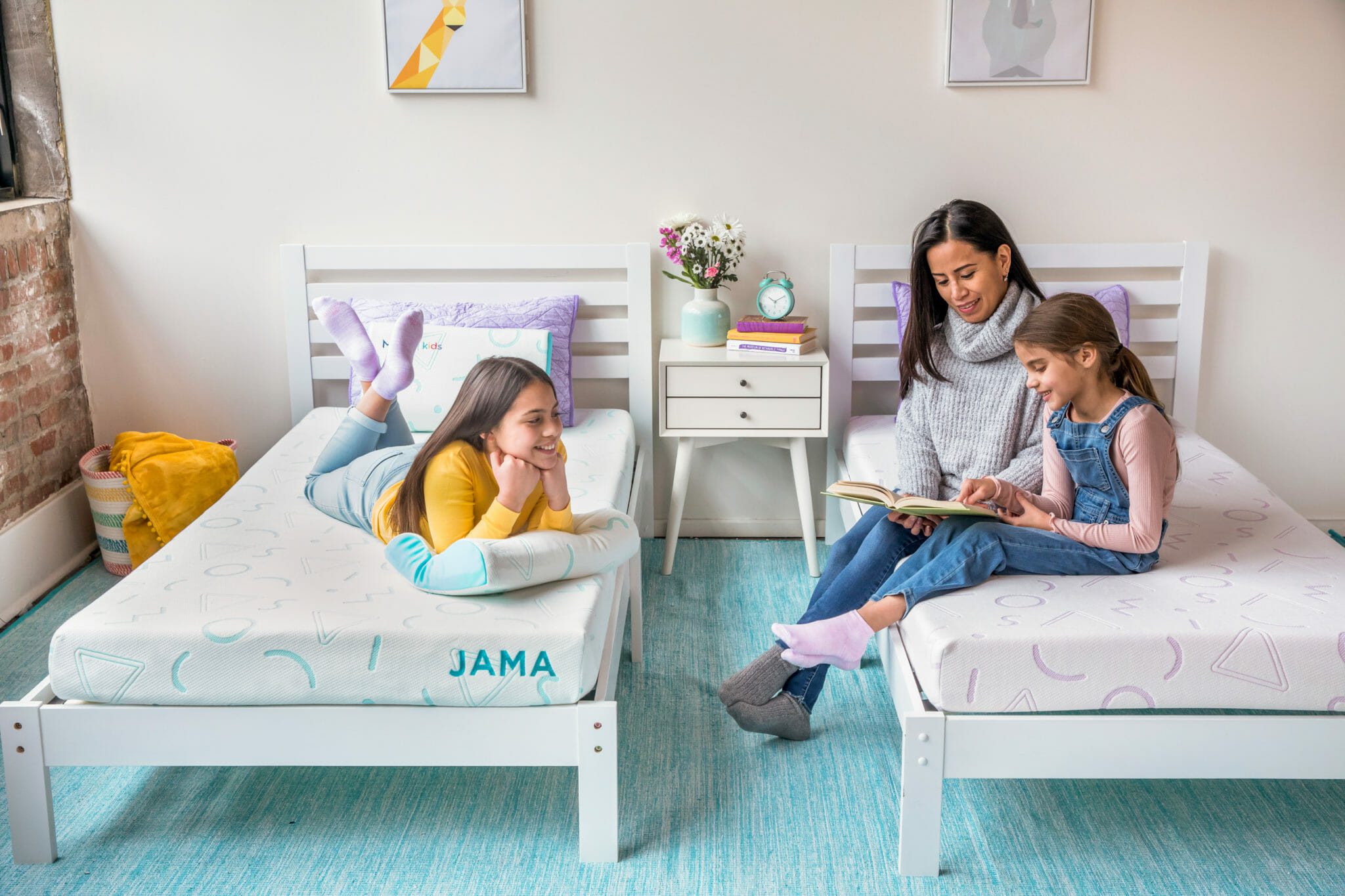 A woman sits on a bed reading a book to two children in a brightly lit room. Both children are lying on their Mlily JAMA 5" Mattresses, attentively listening. The beds and room decor are in light pastel colors, creating a cheerful and cozy atmosphere.