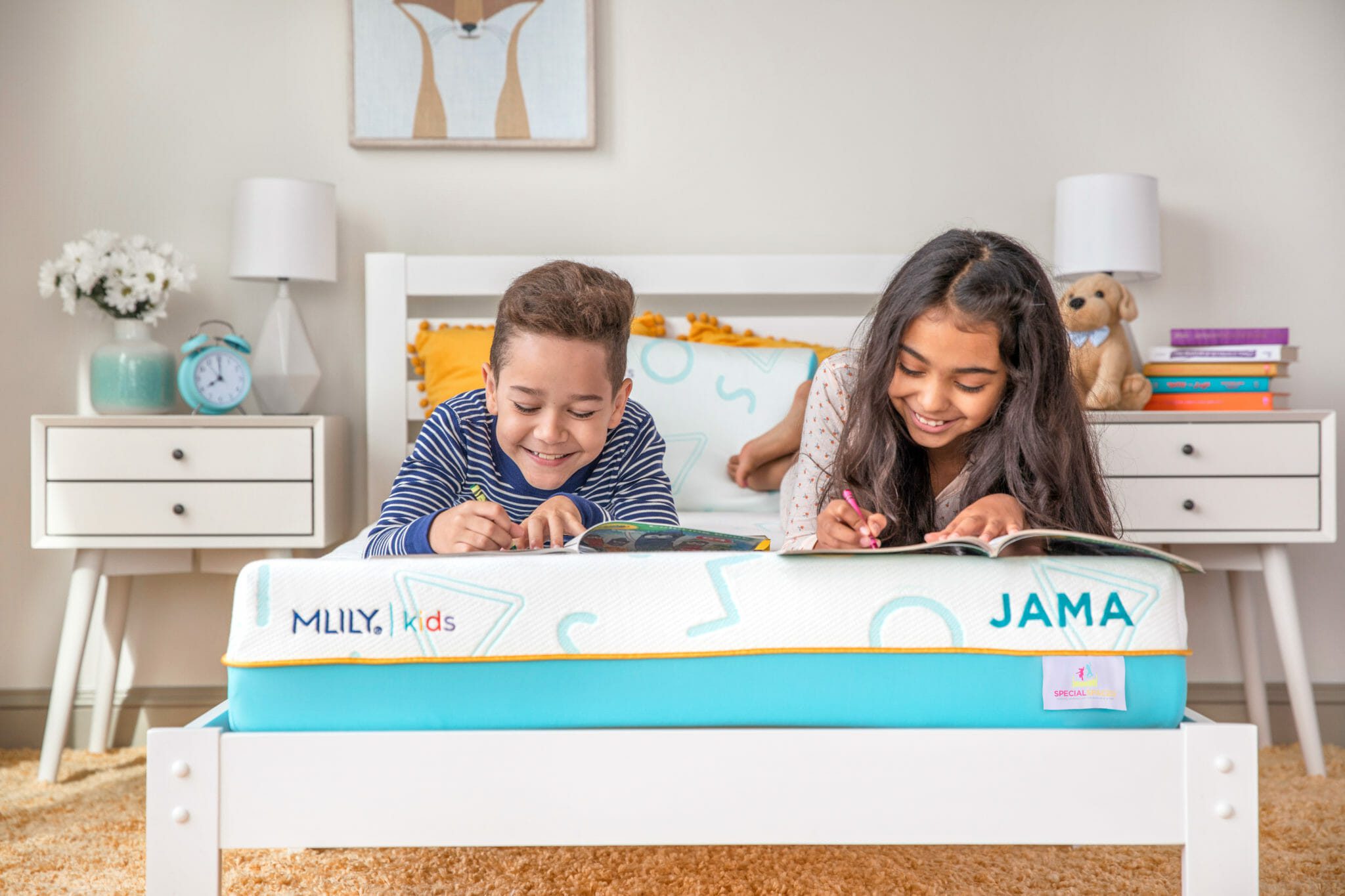 Two children are lying on their stomachs on a bed, happily engaged in reading. The bed features a bright blue and white mattress labeled "Mlily JAMA 7" Mattress." The room is decorated with nightstands, a clock, a teddy bear, books, and wall art.
