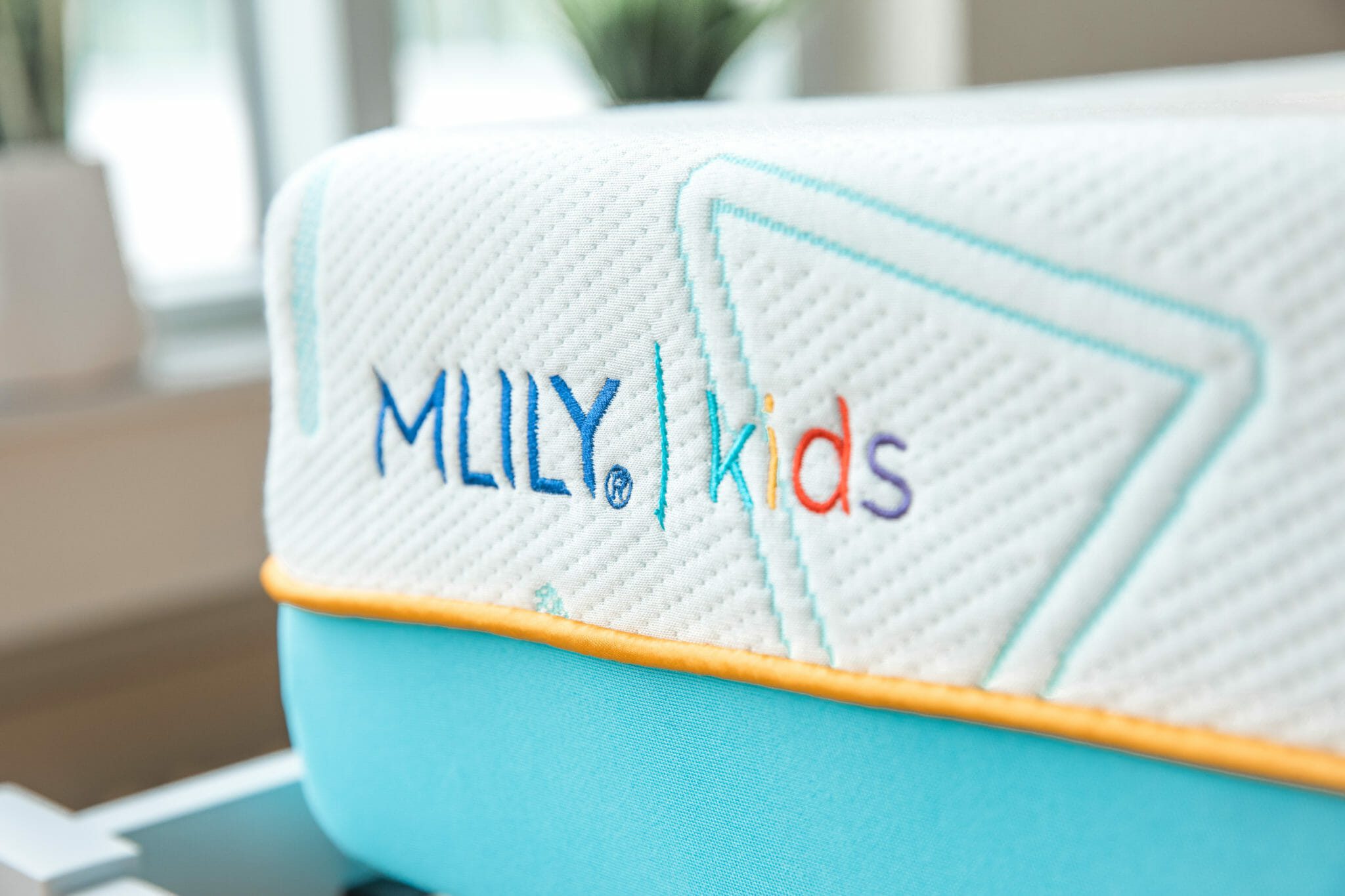 Close-up of a children's mattress with a white top and light blue sides, bordered with a yellow strip. The top of the Mlily JAMA 7" Mattress features the embroidered text "MLILY kids" in playful, colorful letters with geometric stitching patterns.
