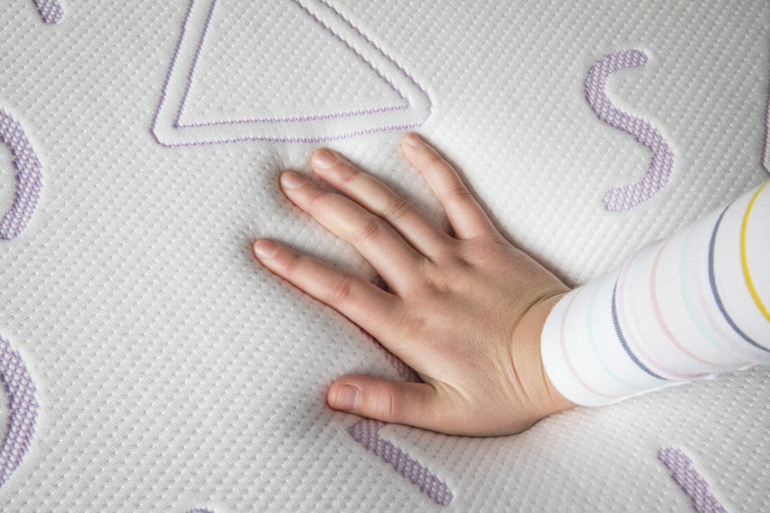 A hand wearing a sleeve with colorful stripes presses down on the Mlily JAMA 7" Mattress, which features textured, raised patterns and symbols. The mattress surface appears soft and responsive to the touch.