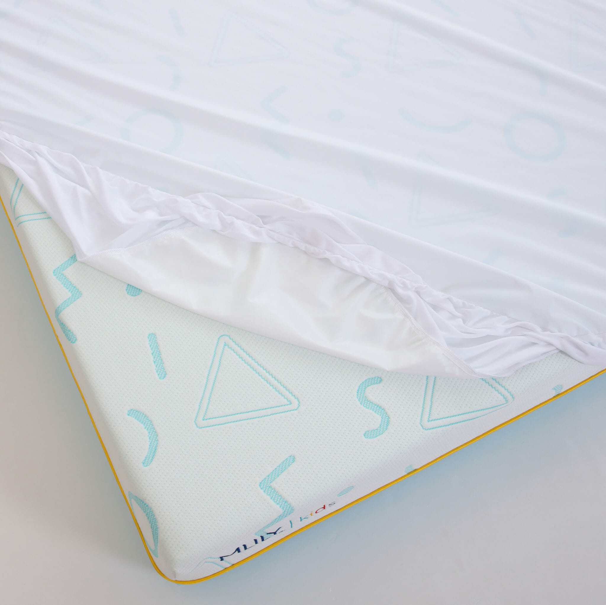 A close-up of the Mlily JAMA 7" Mattress with a corner of a white fitted sheet partially pulled back, revealing the patterned cover beneath. The mattress cover features various geometric shapes in light blue, and the edge is outlined in yellow.
