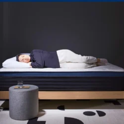 A person is lying on their side on a neatly made Helix Midnight Luxe bed with a dark frame, dressed in a dark hoodie and light pants. The room has dark walls, and a small grey ottoman with a glass of water on it is placed near the bed, resting on a patterned light rug.