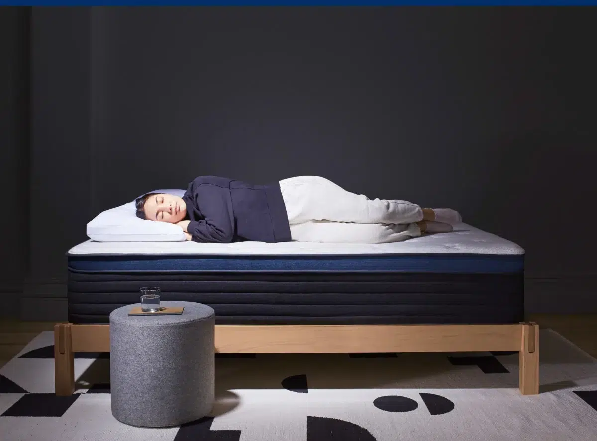 A person is lying on their side on a neatly made Helix Midnight Luxe bed with a dark frame, dressed in a dark hoodie and light pants. The room has dark walls, and a small grey ottoman with a glass of water on it is placed near the bed, resting on a patterned light rug.