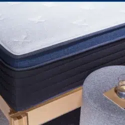 A close-up view shows the Helix Midnight Luxe, a neatly made bed with a plush mattress featuring a white top layer and dark blue sides. Beside the bed is a gray upholstered round ottoman adorned with a gold tray holding a glass of water. The bed frame is crafted from light-colored wood.