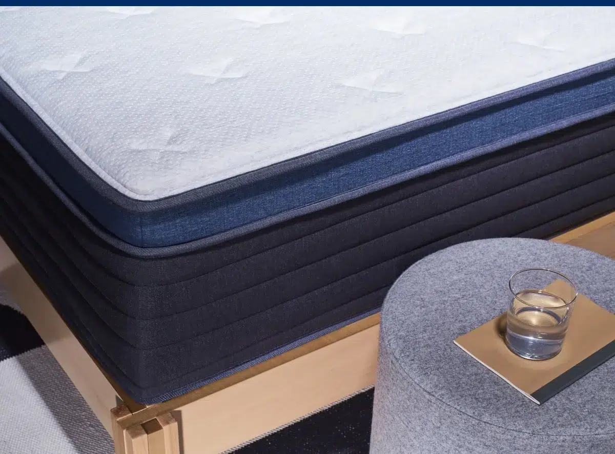 A close-up view shows the Helix Midnight Luxe, a neatly made bed with a plush mattress featuring a white top layer and dark blue sides. Beside the bed is a gray upholstered round ottoman adorned with a gold tray holding a glass of water. The bed frame is crafted from light-colored wood.