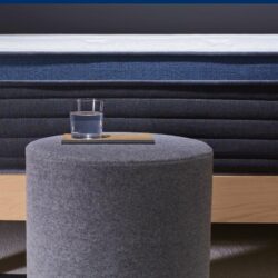 A close-up view of a neatly made bed featuring the Helix Midnight Luxe mattress, elegantly draped in dark blue. The bed is supported by a light wooden frame. In front of the bed sits a cylindrical, gray upholstered ottoman with a glass of water carefully placed on top. The background showcases a sophisticated dark wall.