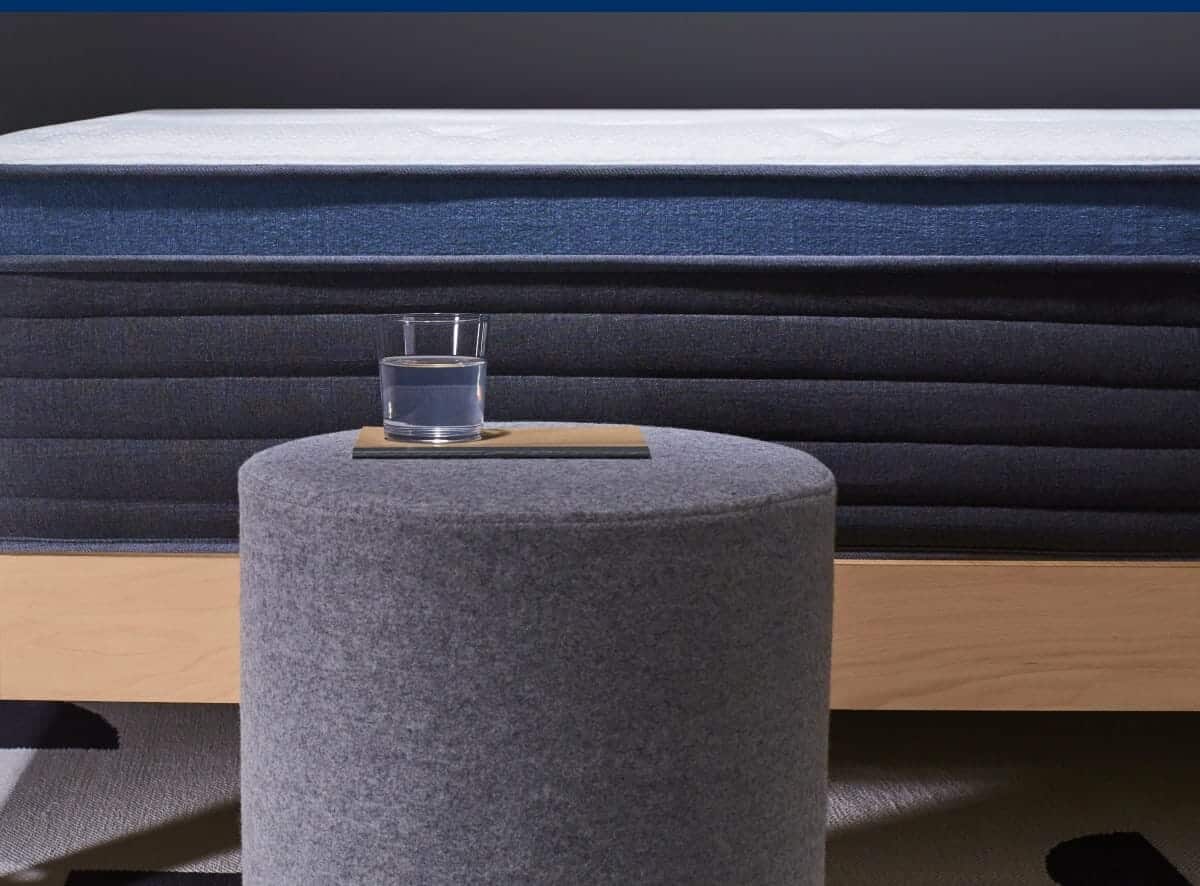 A close-up view of a neatly made bed featuring the Helix Midnight Luxe mattress, elegantly draped in dark blue. The bed is supported by a light wooden frame. In front of the bed sits a cylindrical, gray upholstered ottoman with a glass of water carefully placed on top. The background showcases a sophisticated dark wall.
