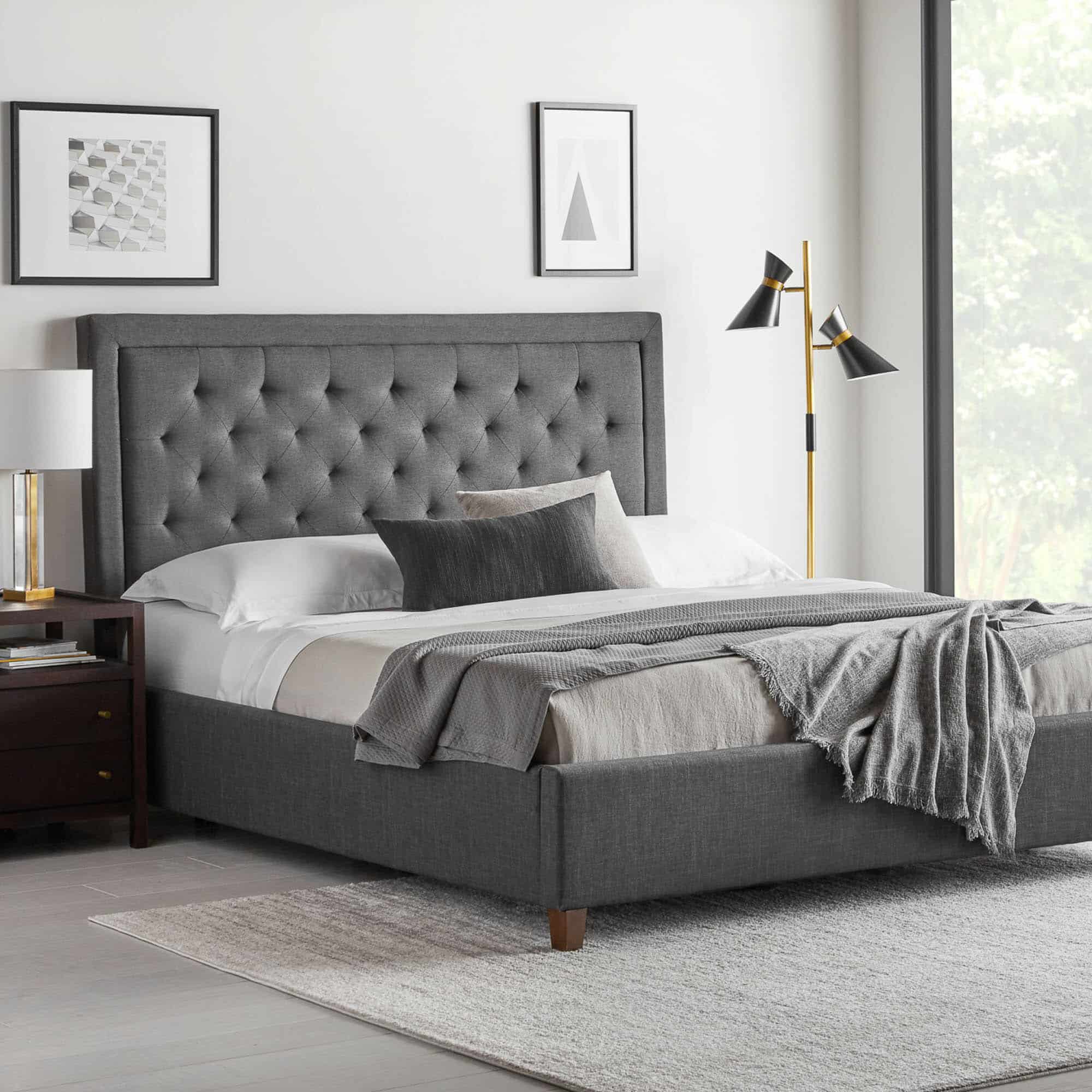 A modern bedroom featuring the large grey Hennessy Headboard bed with white and grey bedding. A dark wood nightstand with a white lamp is on the left, complemented by framed art on the wall above. To the right stands a tall black and gold floor lamp. A light rug covers the floor.