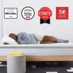 A person relaxes on the Helix Plus, a bed featuring a gray frame and white mattress, dressed in a gray long-sleeve top and shorts. Displayed in the background are four award badges from Wirecutter, Forbes Vetted, CNET, and the American Chiropractic Association.