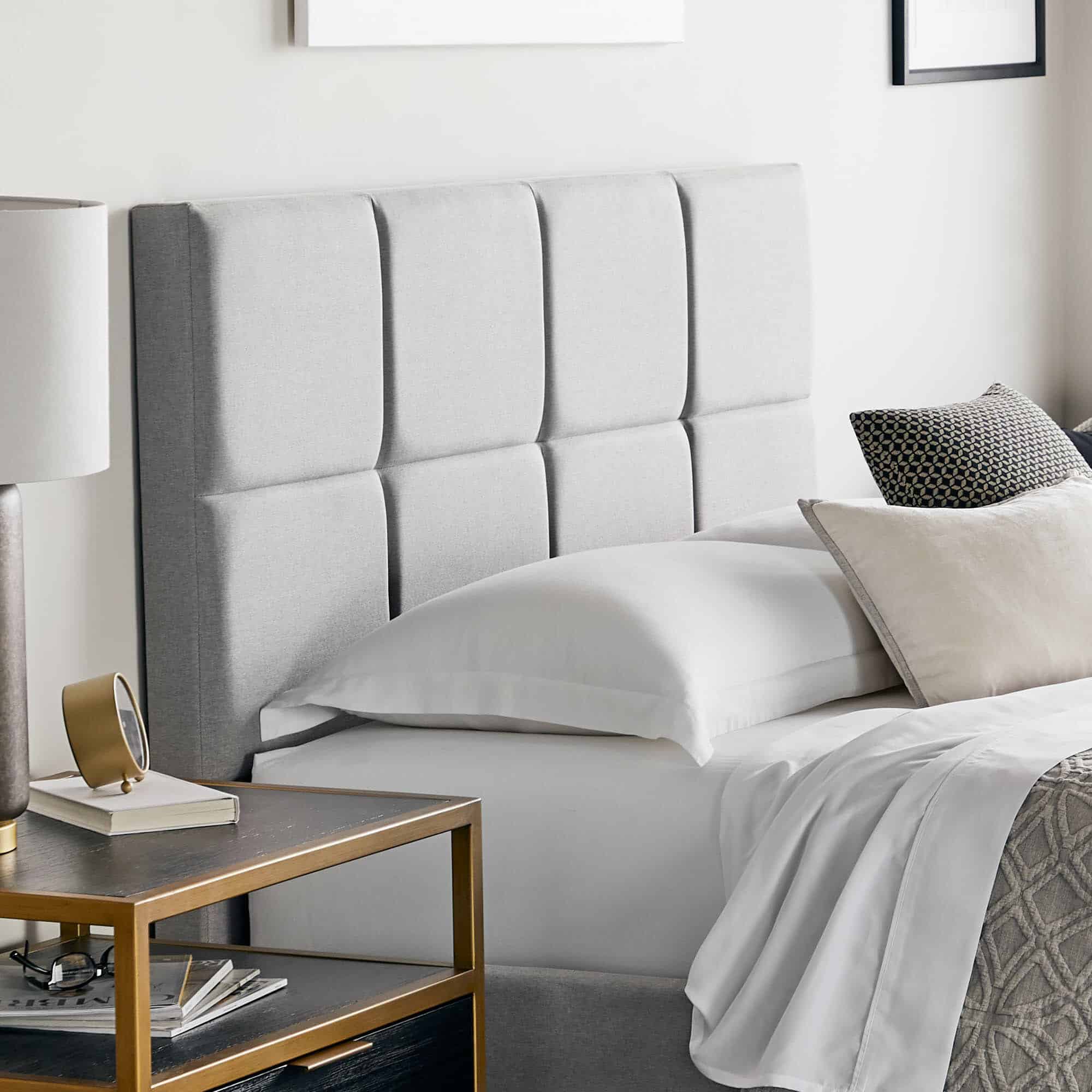 A modern bedroom featuring the Scoresby Designer Bed with a gray upholstered headboard, complemented by white bed linens and a mix of white and gray pillows. A bedside table holds a lamp, books, and small decorative items next to the bed. The walls are decorated with framed artwork.