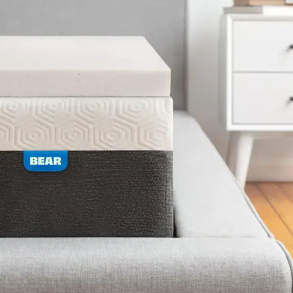 A close-up view of a neatly made bed with a gray upholstered headboard, featuring a Bear Pro Mattress Topper and a mattress labeled "BEAR." In the background, there's a white nightstand with two drawers and a book on top, set against a light-colored wall. The floor is wooden.
