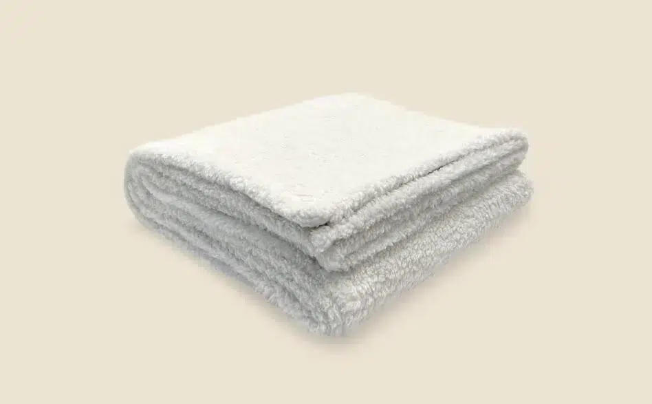 A neatly folded Puffy Deluxe Blanket, in a light grey hue, is set against a plain beige background. The texture of the blanket appears soft and plush, suggesting it's warm and comfortable.
