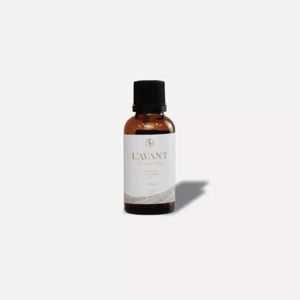 A small amber glass bottle with a black cap is placed against a plain white background. The label reads "L'AVANT Collective" with additional text underneath, including the product name "Fresh Linen Laundry Oil." The bottle casts a soft shadow to the right.