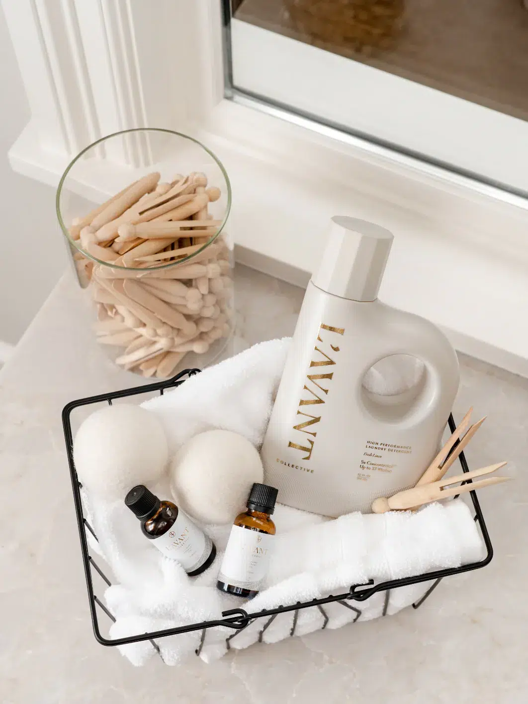 A stylish bathroom setup featuring a glass jar filled with reusable wooden cotton swabs, a black wire basket containing a white towel, two small essential oil bottles, a bottle of Fresh Linen Laundry Oil by L'AVANT Collective, two wool dryer balls, and wooden clothespins.