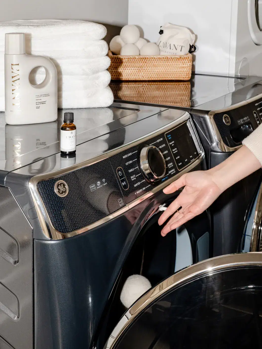A person is opening the door of a front-loading washing machine in a laundry room. Various laundry products, including folded white towels, detergent, and a small bottle of Fresh Linen Laundry Oil from L'AVANT Collective, are neatly arranged on top of the machines.