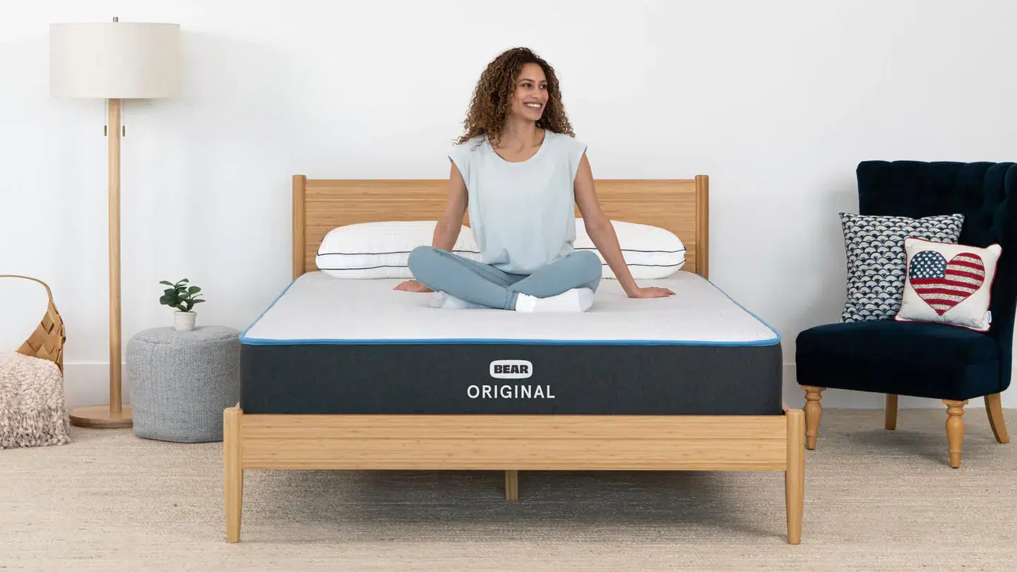 A woman with curly hair and casual attire sits cross-legged on a neatly made bed featuring a Bear Original mattress. The room includes a wooden bed frame, a floor lamp, a plant, a woven basket, and a navy armchair adorned with an American flag cushion.