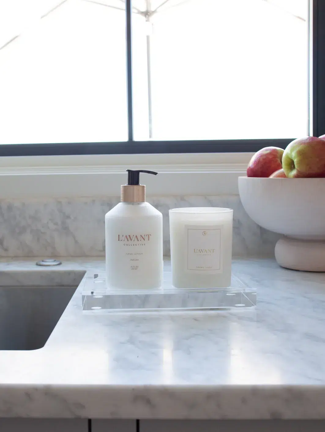 A marble counter beside a stainless steel sink features a Lucite Tray - Medium by L'AVANT Collective, elegantly holding hand soap and a candle in white containers with gold accents. In the background, a bowl filled with apples sits on the counter near a window.