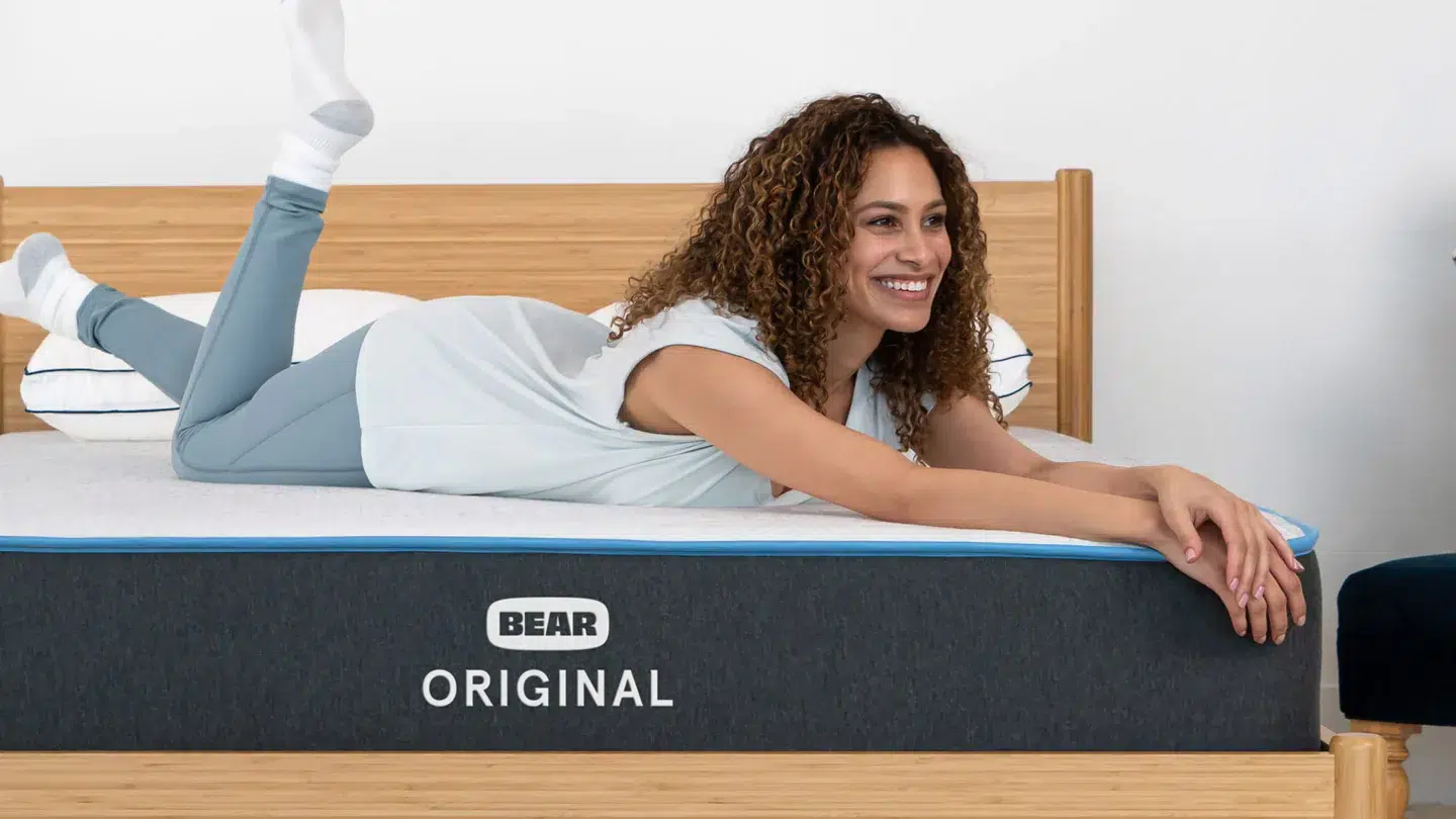 A woman with curly hair, dressed in a light blue sleeveless top and matching light blue pants, is reclining on her stomach on a Bear Original mattress placed atop a wooden bed frame. She smiles and gazes off to the side, with her legs bent at the knees and feet raised up.