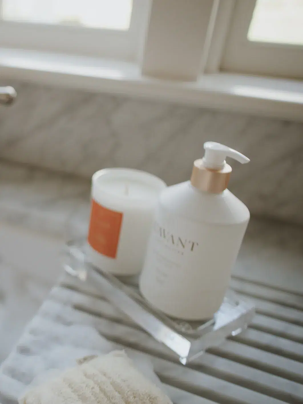 A Lucite tray from L'AVANT Collective on a countertop next to a window holds a white soap dispenser with an elegant pump and a candle in a white jar with an orange label. In the foreground, there is a folded towel.