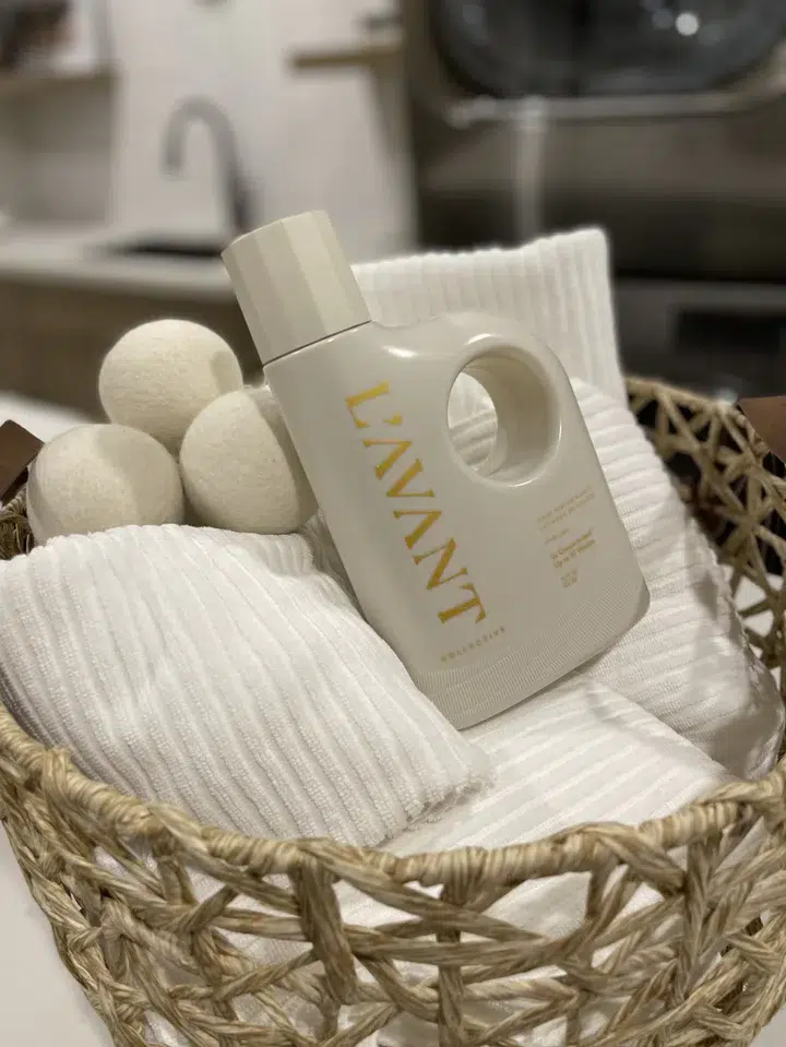 A beige woven basket contains white ribbed towels, three wool dryer balls, and a white bottle of L'AVANT High Performing Laundry Detergent - Fresh Linen. The setting appears to be a laundry room with a blurred background featuring a sink and counter.