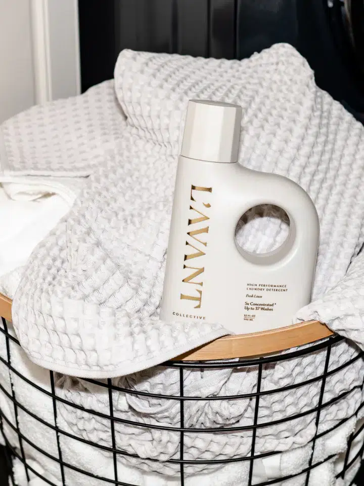 A black wire laundry basket filled with folded white and waffle-textured towels. A bottle of L'AVANT High Performing Laundry Detergent - Fresh Linen rests on top, featuring a modern design with a handle and gold lettering. The scene is well-lit, highlighting the product.