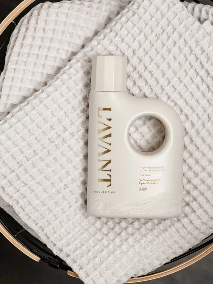 A bottle of L'AVANT High Performing Laundry Detergent - Fresh Linen sits on a white waffle-textured towel in a black basket. The bottle is a matte off-white color with gold lettering and features a distinctive round handle integrated into the design.