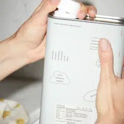 A person holds a metal container labeled as Dedtergent 05 "Spring," adorned with a white screw cap, printed text, and cloud designs. The text specifies that it is a gentle laundry detergent for infants. In the background, fabric with a yellow smiley face pattern is partially visible.