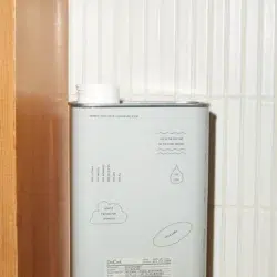 A large, rectangular container labeled "Dedtergent 05 'Spring'" with a white lid is placed on a wooden shelf. The container is light gray with minimalist text and graphics, featuring phrases like "Direction your life in a singular energy" along with illustrations of clouds and a heart.