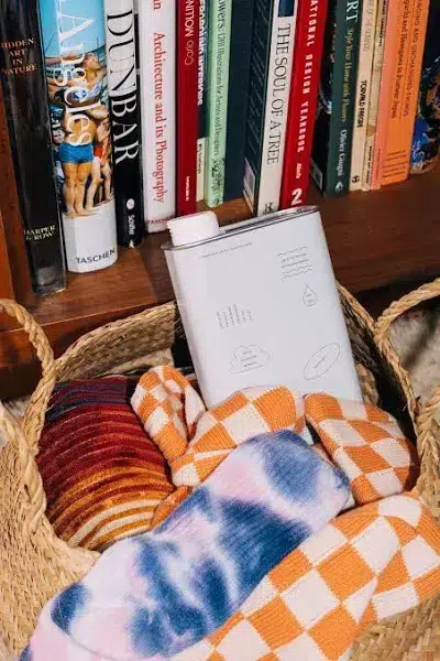 A woven basket containing colorful striped and checkered clothing items, with a white Dedtergent 01 "Taunt" product box placed on top. The basket is set against a background of a bookshelf filled with various books on architecture and art.