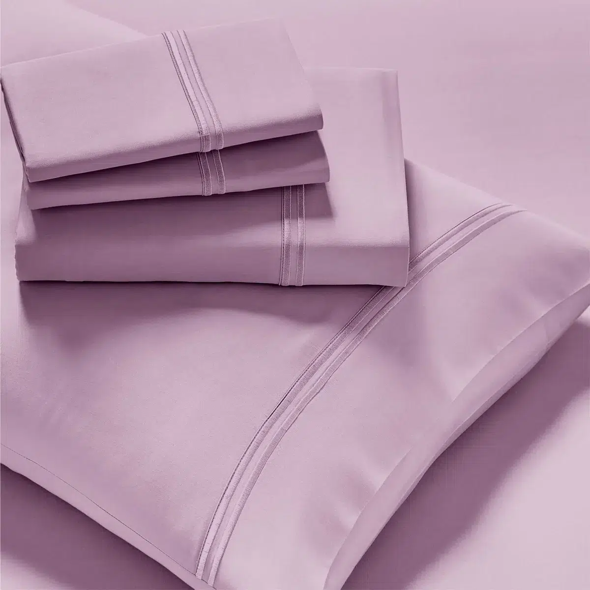 A neatly arranged stack of pastel-colored Refreshing TENCEL™ Lyocell Sheets - PureCare, including pillowcases, sheets, and a duvet cover, displayed against a matching background. The linens feature simple, elegant stitch detailing for a clean, sophisticated look.