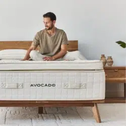 A man sits on the edge of a bed in a minimalist bedroom. The bed has an Avocado Luxury Mattress atop a wooden frame. The man, wearing a beige t-shirt and white pants, smiles while touching the bed. The room features neutral decor with ceramic vases on a wooden nightstand.