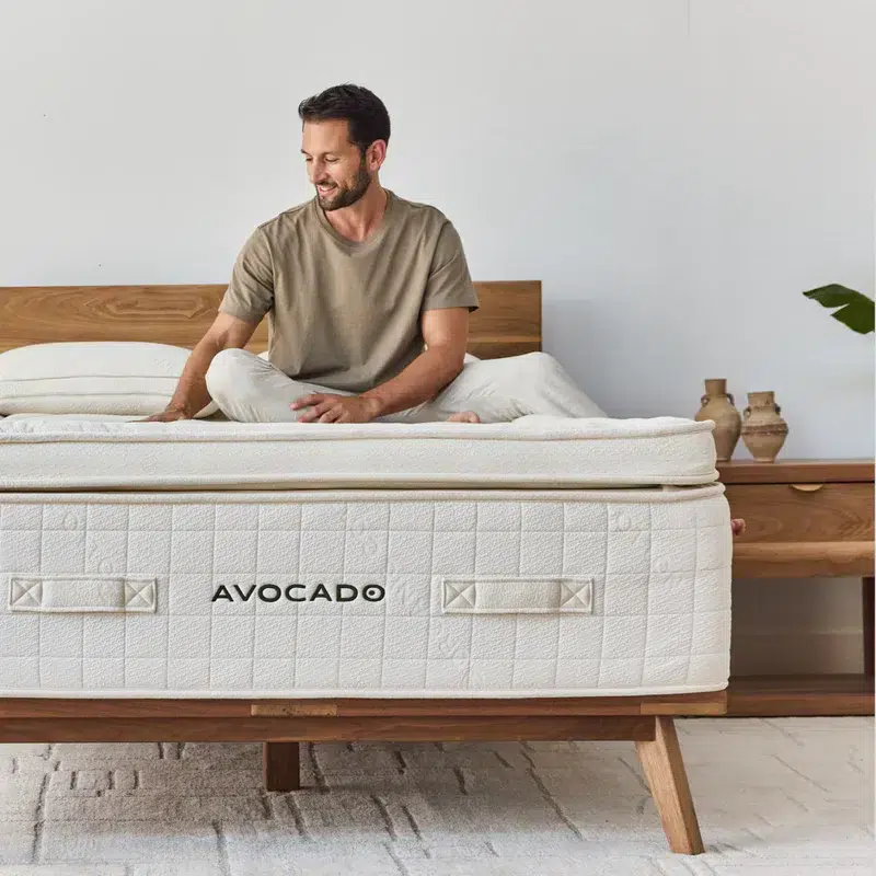 A man sits on the edge of a bed in a minimalist bedroom. The bed has an Avocado Luxury Mattress atop a wooden frame. The man, wearing a beige t-shirt and white pants, smiles while touching the bed. The room features neutral decor with ceramic vases on a wooden nightstand.