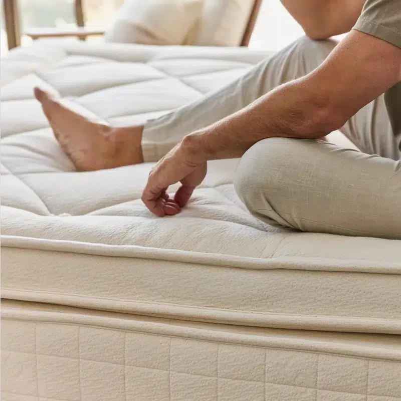 A person is sitting on the edge of an Avocado Luxury Mattress, pressing down with their hand to test its firmness. The mattress features a quilted design with a soft, cushioned appearance, and the person is wearing light-colored pants. The room appears bright and airy.