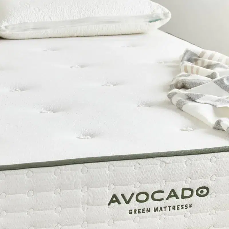A close-up of a neatly made Avocado Green Mattress showcases its white surface adorned with a subtle green trim. The brand name "AVOCADO" and "GREEN MATTRESS" are printed on its side. A pillow and a striped blanket lie atop the mattress, adding a cozy touch.