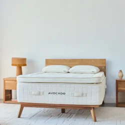 A minimalist bedroom features a wooden bed frame with an Avocado Luxury Mattress. The bed is flanked by two wooden nightstands, one with a lamp and the other with a small decorative object. The room has white walls and a light-colored rug on the floor.