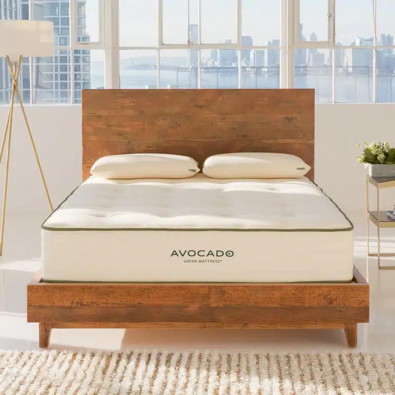 In a modern bedroom, an impeccably arranged bed showcasing the Avocado Green Mattress takes center stage. The mattress, draped with two plush pillows, sits elegantly on an eco-friendly wooden frame. Expansive windows in the background provide a stunning view of the city skyline and bathe the room in natural light.