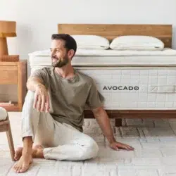 A man sits on the floor beside a bed with an "Avocado Luxury Mattress." He is looking away and smiling, wearing a green t-shirt and white pants. The bedroom features a wooden headboard, nightstand with a lamp, and exudes a cozy, bright atmosphere.
