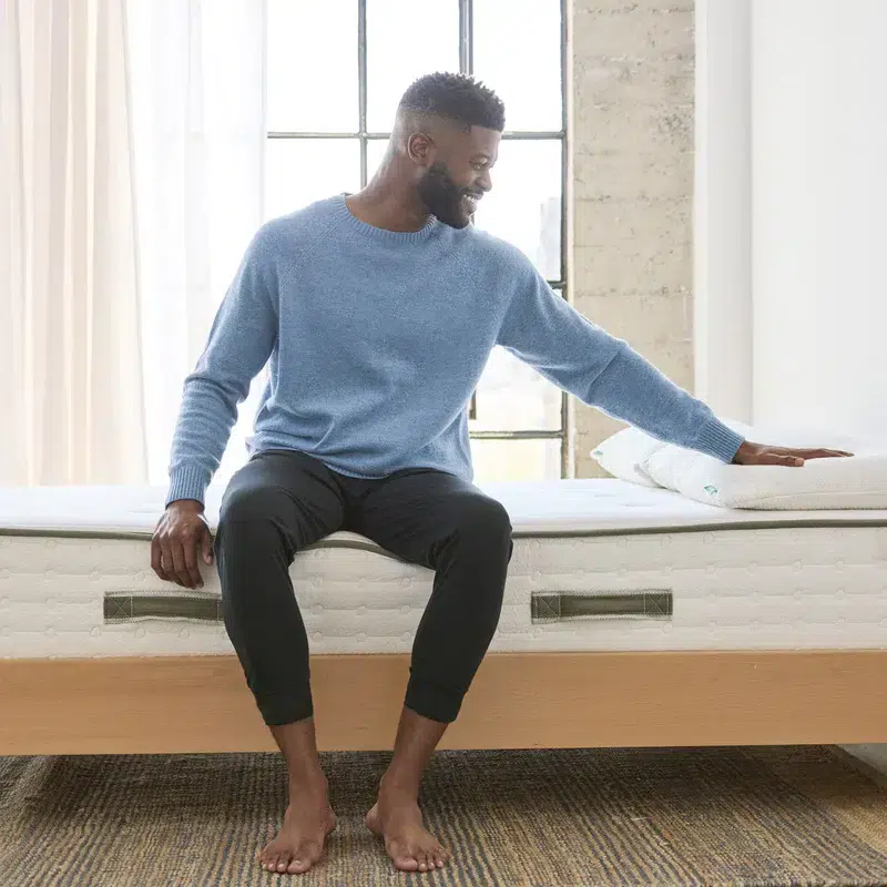 A man with short hair and a beard, wearing a blue sweater and dark pants, sits barefoot on the edge of an Avocado Green Mattress in a minimalist, well-lit room with a brick wall and large window in the background. He is smiling and looking at his hand, which rests on a pillow.