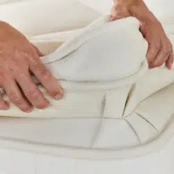 Close-up of a person adjusting the Avocado Luxury Mattress cover. The person’s hands are lifting a corner of the off-white, plush fabric, revealing the layered structure of the mattress beneath. The material appears soft and cushioned for comfort.