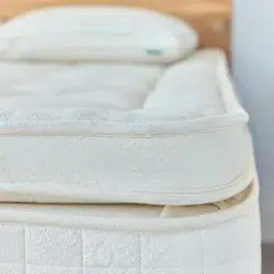 A close-up of a neatly made bed featuring the corner of an Avocado Luxury Mattress adorned with a thick, plush mattress topper. The white topper adds extra comfort to the bed. A pillow is visible at the head of the bed in the background.