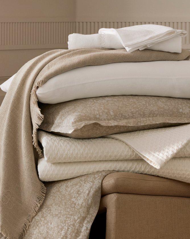 A neatly folded stack of beige and white bed linens is positioned on a beige ottoman. The linens include a variety of textured blankets, sheets, and pillowcases. The calming, neutral colors add to a serene and cozy atmosphere.