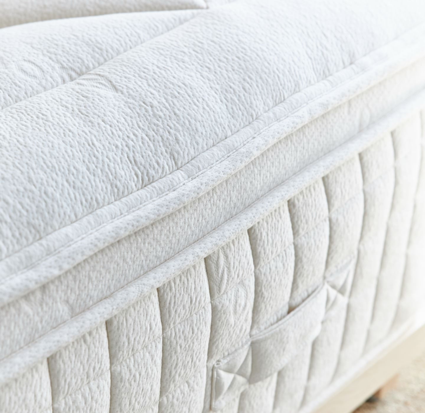 Close-up view of the corner of a white, plush, quilted mattress. The mattress features a textured fabric with horizontal and vertical seams, and includes a small fabric loop for easy rotation. The image highlights the soft and cushioned surface of the mattress.