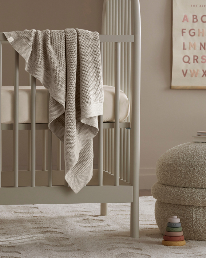 A cozy nursery scene featuring a gray crib with a beige knitted blanket draped over it. A soft ottoman and a stackable toy are on a textured rug nearby. In the background, a wall poster displays the alphabet in a minimalist style.