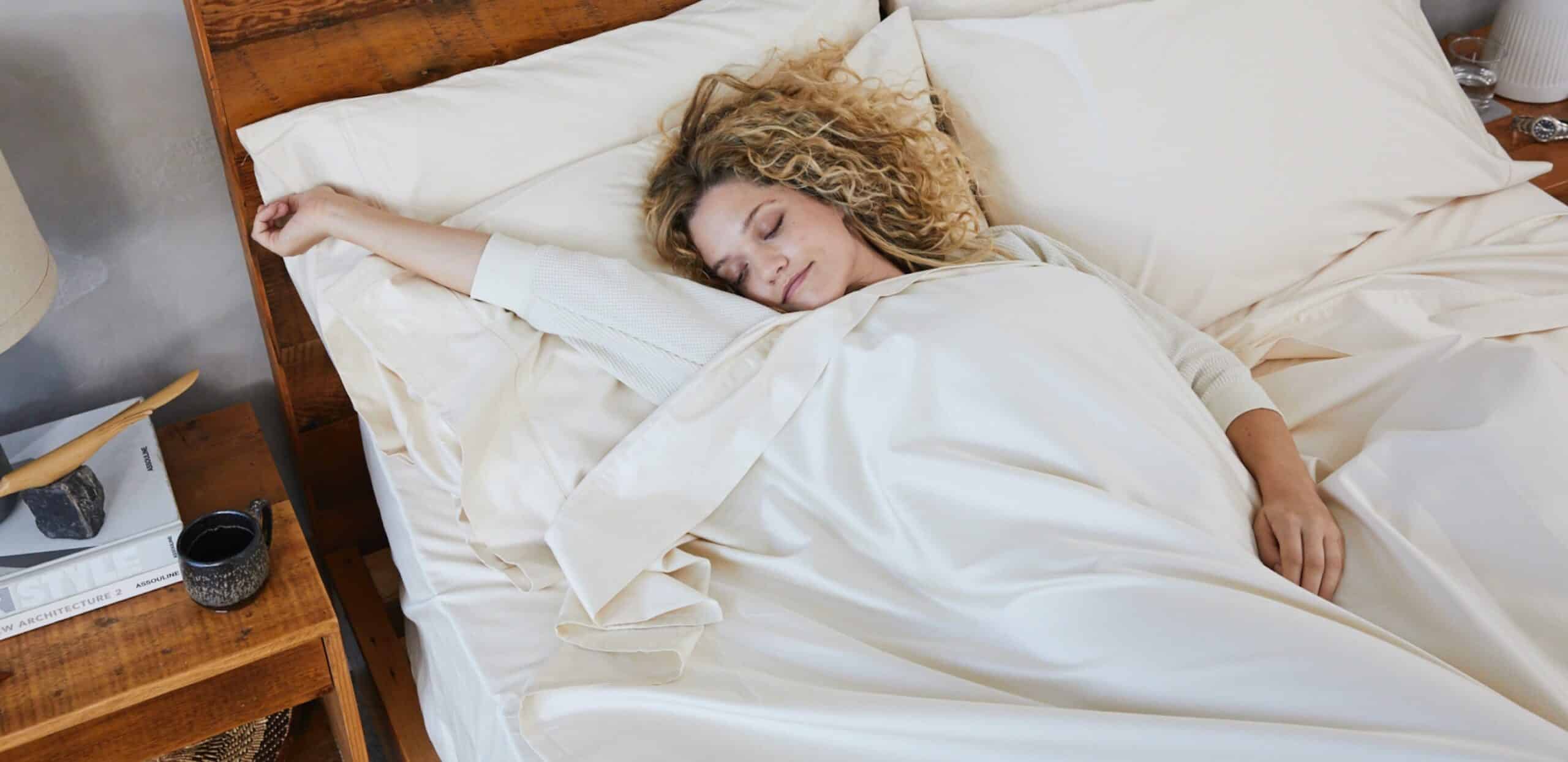 A person with curly hair is lying in bed, covered by cream-colored sheets, with one arm stretched out on a wooden headboard. A small bedside table with a mug, a book, and a lamp is visible next to the bed. The person appears to be peacefully sleeping.