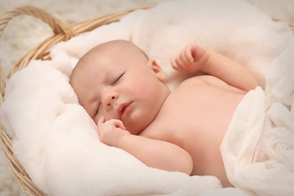 A sleeping baby lies on its back in a wicker basket lined with soft, white blankets. The baby is peacefully resting with one hand near its face. The background is softly blurred, emphasizing the soothing and cozy atmosphere.