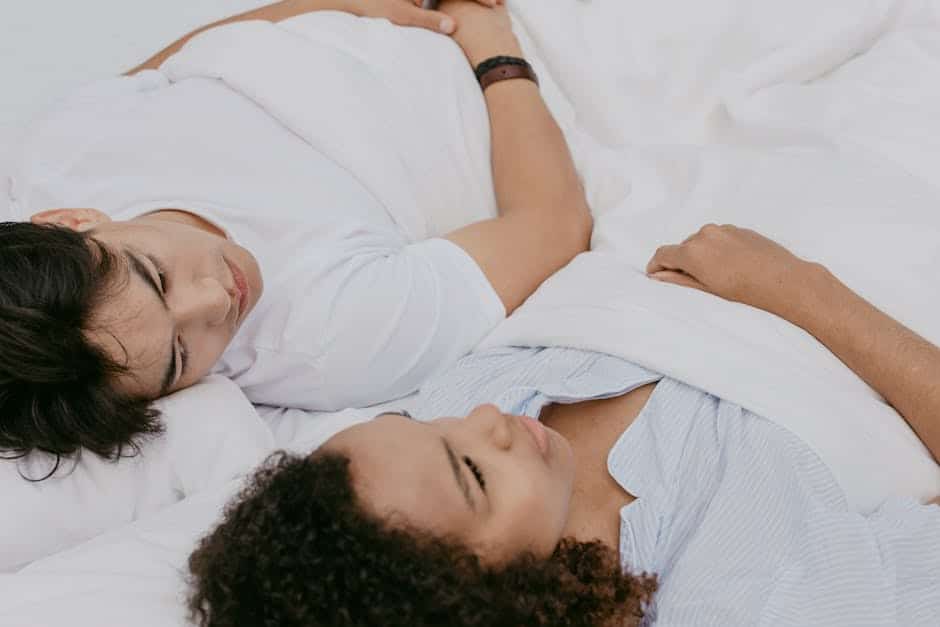 Two people are lying together in bed, looking at each other under a white blanket. The person on the left is wearing a white shirt, while the person on the right is wearing a light blue and white striped shirt. Their heads are resting on pillows.
