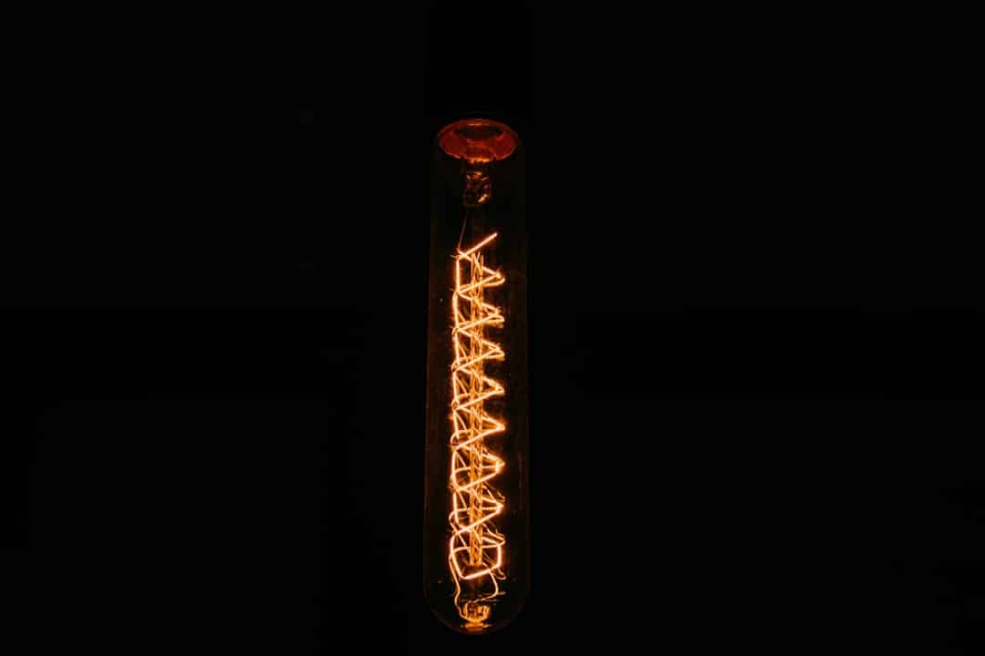 A single vertical light bulb against a dark background, with visible glowing filaments forming a zigzag pattern inside the bulb, casting a warm, orange light.