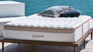 A Loom & Leaf mattress, known for its Saatva contour 5 support, is placed on a wooden bed frame outdoors, with a body of water visible in the background. Two pillows, one gray and the other blue, lay on top of the mattress. The setting is bright and serene.