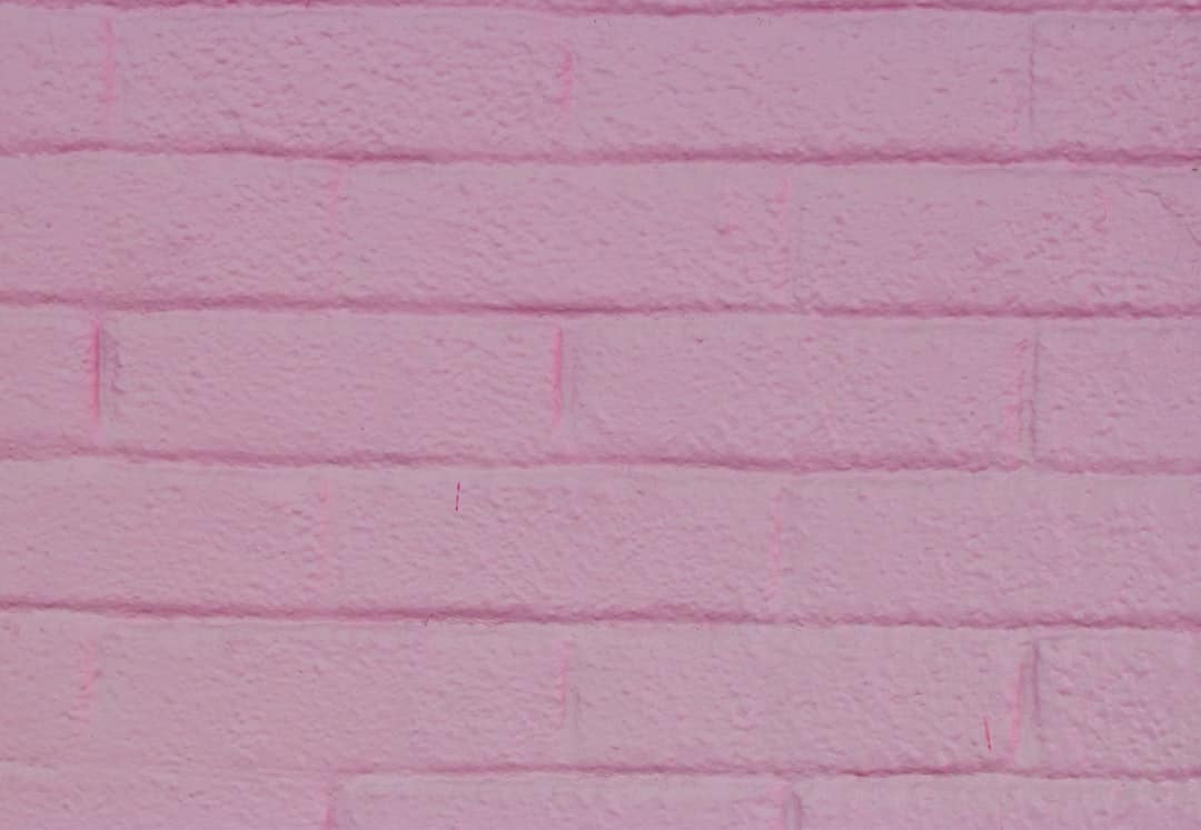 A close-up of a textured brick wall painted in a soft pink color. Each brick has visible grooves and rough surfaces, giving the wall a slightly uneven, tactile appearance. The overall effect is a vibrant and cheerful backdrop.