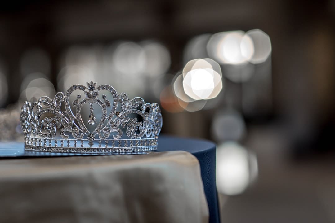 Close-up of an ornate silver tiara decorated with intricate patterns and gemstones, resting on a table with a blurred background of soft lights. The tiara has a heart-shaped design at its center, creating an elegant and delicate appearance.